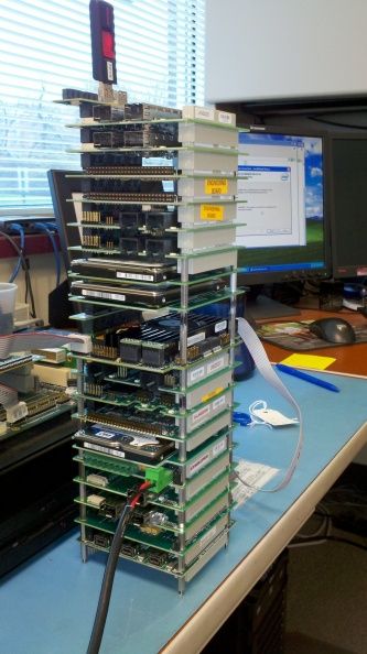 Tall stack of green PCB boards on a desk, with monitors, cables, and office items in the background