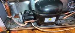 Widok na agregat lodówki Beko z widocznym czarnym plastikowym elementem i białym cylindrycznym kondensatorem obok.