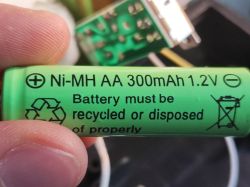Ręka trzymająca zielony akumulator Ni-MH AA o pojemności 300mAh i napięciu 1,2V.