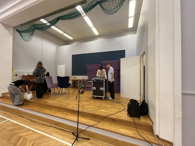 Grupa osób przygotowuje sprzęt nagłośnieniowy na scenie w sali gimnastycznej.
