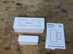 Sonoff DW2-Wi-Fi Door Sensor Set Sonoff DW2-Wi-Fi sensor set with user guide on a wooden table.