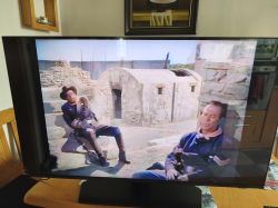 Telewizor na drewnianym stole wyświetla western; w tle wnętrze kuchni