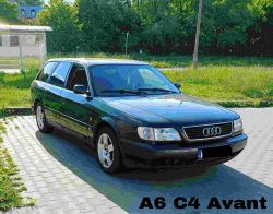 Ciemny samochód Audi A6 C4 Avant zaparkowany na bruku, z zielonymi drzewami w tle.