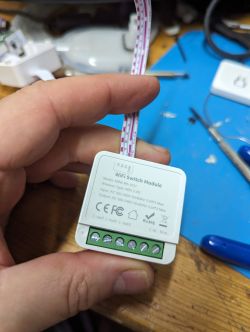 A hand holding a WiFi switch module with long wires, against a workbench background.
