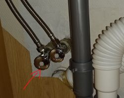 Rury wodociągowe i odpływowe z metalowymi i plastikowymi złączami, z czerwonymi strzałkami wskazującymi na zawór.