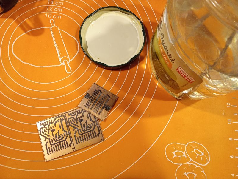 The image shows a kitchen mat with drawn circles, a jar labeled Ogórki kwaszone, a lid, and two printed circuit boards.