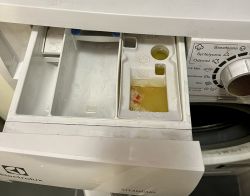 Szuflada pralki Electrolux z zalegającą żółtą wodą i resztką kapsułki w przegródce na płyn zmiękczający.