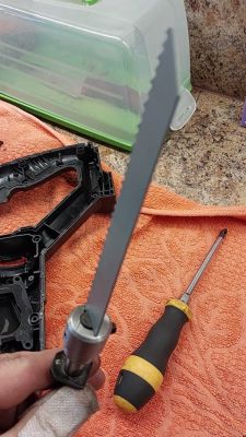 Ręka w rękawiczce trzyma piłę z brzeszczotem, obok leży śrubokręt i plastikowa rama na pomarańczowym ręczniku.