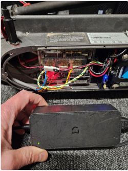 Ręka trzymająca ładowarkę podłączoną do wnętrza elektrycznej hulajnogi z widocznymi kablami i komponentami.