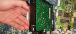 Dłoń obok zielonej płytki PCB w czarnej obudowie, na tle niebieskiego materiału
