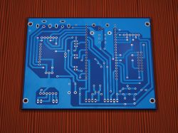Bottom view of a blue PCB with copper traces and many vias on a wood-like textured background