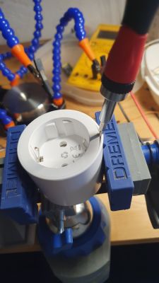 Close-up of a white plug held in a vise, with a screwdriver inserted into its female part.