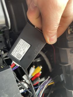 Ręka trzyma moduł elektroniczny w samochodzie z widocznymi przewodami i złączami.