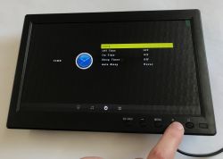 Small black monitor showing “Clock” menu; a finger presses the “+” button on the bottom control panel.