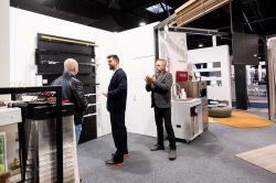 Three men talk at a trade-show booth with a display of drawers and fabric or material samples.