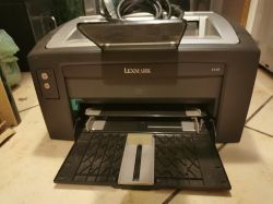 Drukarka Lexmark E120 na podłodze z otwartą tacą na papier i widocznym przewodem zasilającym.