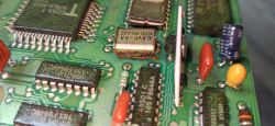 Zbliżenie na płytkę drukowaną z różnymi elementami elektronicznymi, w tym scalakami, kondensatorami i elementem oznaczonym jako EAVF-AA NDK 8641, który wygląda na skorodowany.