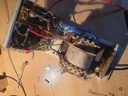 Wnętrze starszego prostownika leży na drewnianym stole, pokazując elementy elektroniczne, przewody i transformator.