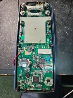Otwarta obudowa urządzenia z zieloną płytką PCB, bezpiecznikami u góry i baterią guzikową