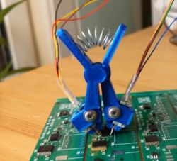 Niebieski, drukowany 3D klips testowy ze sprężynką dociskającą, podłączony przewodami do układu scalonego na płytce PCB.