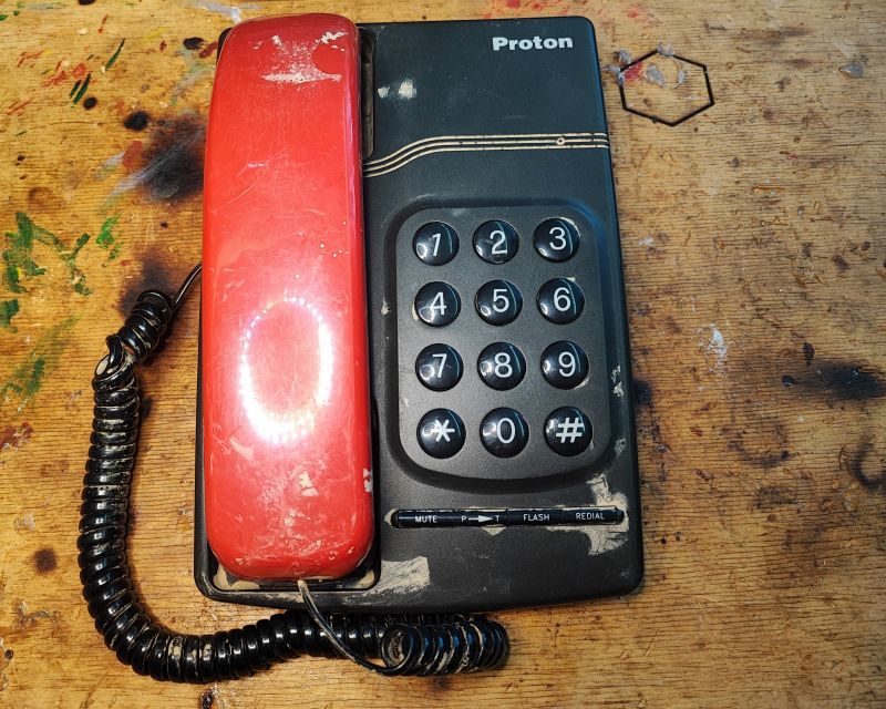 Czerwony telefon stacjonarny Proton T1332 z lat 90. z klawiaturą numeryczną i dodatkowymi przyciskami, na drewnianym blacie.