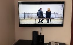 Telewizor wiszący na ścianie wyświetla scenę z filmu, poniżej znajdują się głośniki i gazeta.