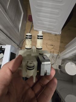 Ręka trzymająca część z urządzenia, prawdopodobnie elektrozawór, na tle pralni.