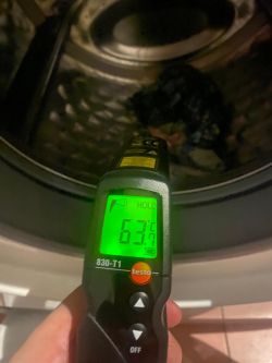 Pirometr przemysłowy pokazujący temperaturę 63,1°C przy drzwiczkach otwartej pralki.