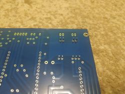 Underside of a blue PCB with through-holes and a mounting hole on a beige textured fabric background