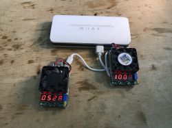Biały powerbank testowany dwoma obciążeniami elektronicznymi z wyświetlaczami i wentylatorami.