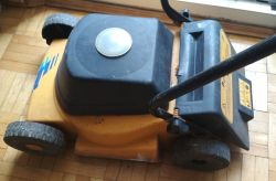 Mała, żółta kosiarka elektryczna z czarną pokrywą na drewnianej podłodze, bez górnej części rączki.