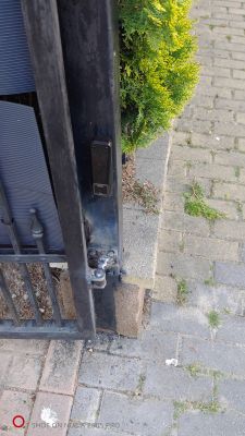 Zbliżenie na fotokomórkę zamontowaną na metalowym słupku bramy wjazdowej, obok kostki brukowej i krzewu.