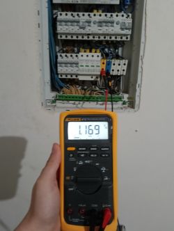 Multimetr pokazujący napięcie 116.9 V przed otwartą skrzynką elektryczną.