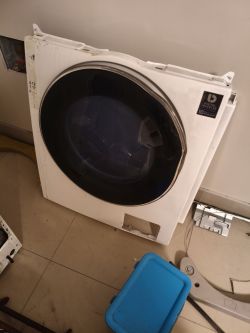 Odłączony przedni panel pralki Samsung z dużymi drzwiami na tle jasnej ściany.