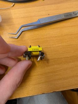 Switching transformer held between fingers on a wooden desk, tweezers nearby.
