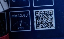 Zbliżenie na wskaźnik akumulatora z grafiką „min 12.4 V” i kodem QR