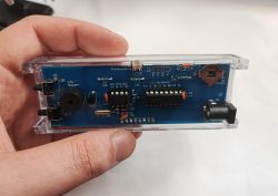 DIY clock PCB in a clear acrylic case held in a hand; visible buttons, buzzer, and DC barrel power jack.