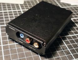 Czarny aluminiowy DAC na macie w kratkę, z gniazdem optycznym i wyjściami RCA oraz jack 3,5 mm