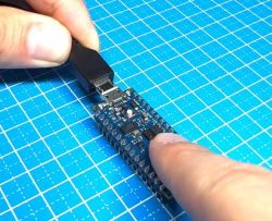 Dłoń podłącza kabel USB do płytki Arduino Nano 33 BLE Sense Lite na niebieskiej kratkowanej macie.