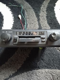 Stare radio samochodowe z analogową skalą i dwoma pokrętłami, leżące na dywanie, z widocznymi luźnymi przewodami.