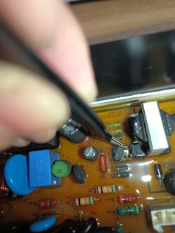 Płytka drukowana z uszkodzonym elementem elektronicznym wskazywanym pęsetą.