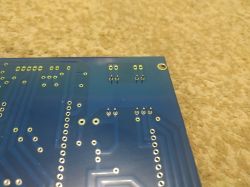 Underside of a blue PCB with plated through-holes and a mounting hole on a beige surface