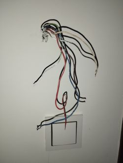 Kilka kolorowych, odsłoniętych przewodów elektrycznych wystaje ze ściany nad białym włącznikiem światła.
