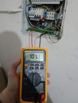Ręka trzymająca miernik napięcia, pokazujący odczyt 101,1 V, na tle otwartej skrzynki elektrycznej.