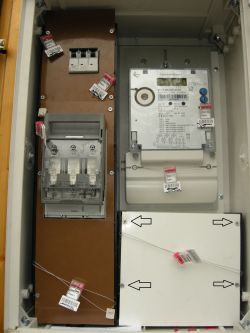 Zbliżenie na licznik elektryczny z plombami i białą osłoną oznaczoną czarnymi strzałkami.