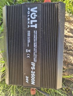Przetwornica napięcia VOLT Polska IPS-2600N 24V leżąca na trawie, widoczny opis parametrów technicznych na obudowie.