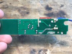 Bottom side of a PCB with visible traces and solder points, held above a wood surface
